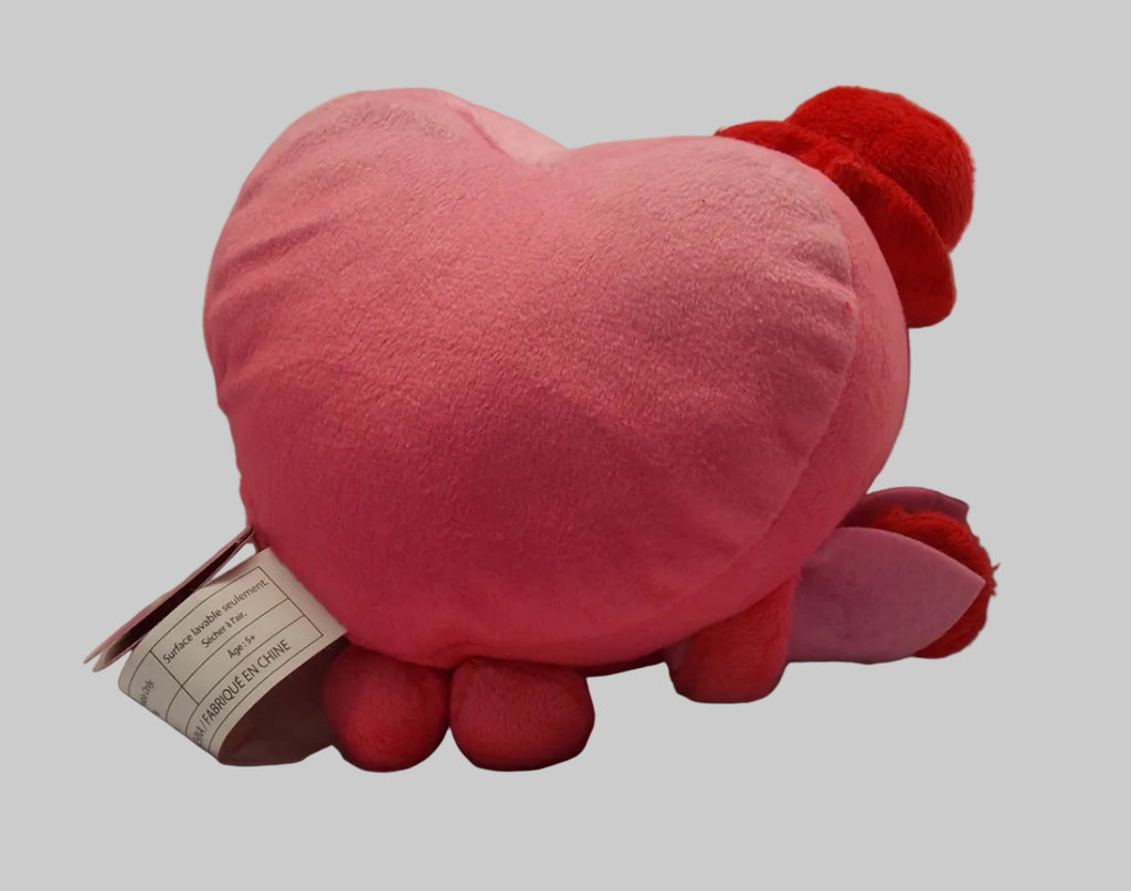 A pink heart-shaped plush pillow with red accents and a white product tag attached, photographed against a wrinkled white fabric background.