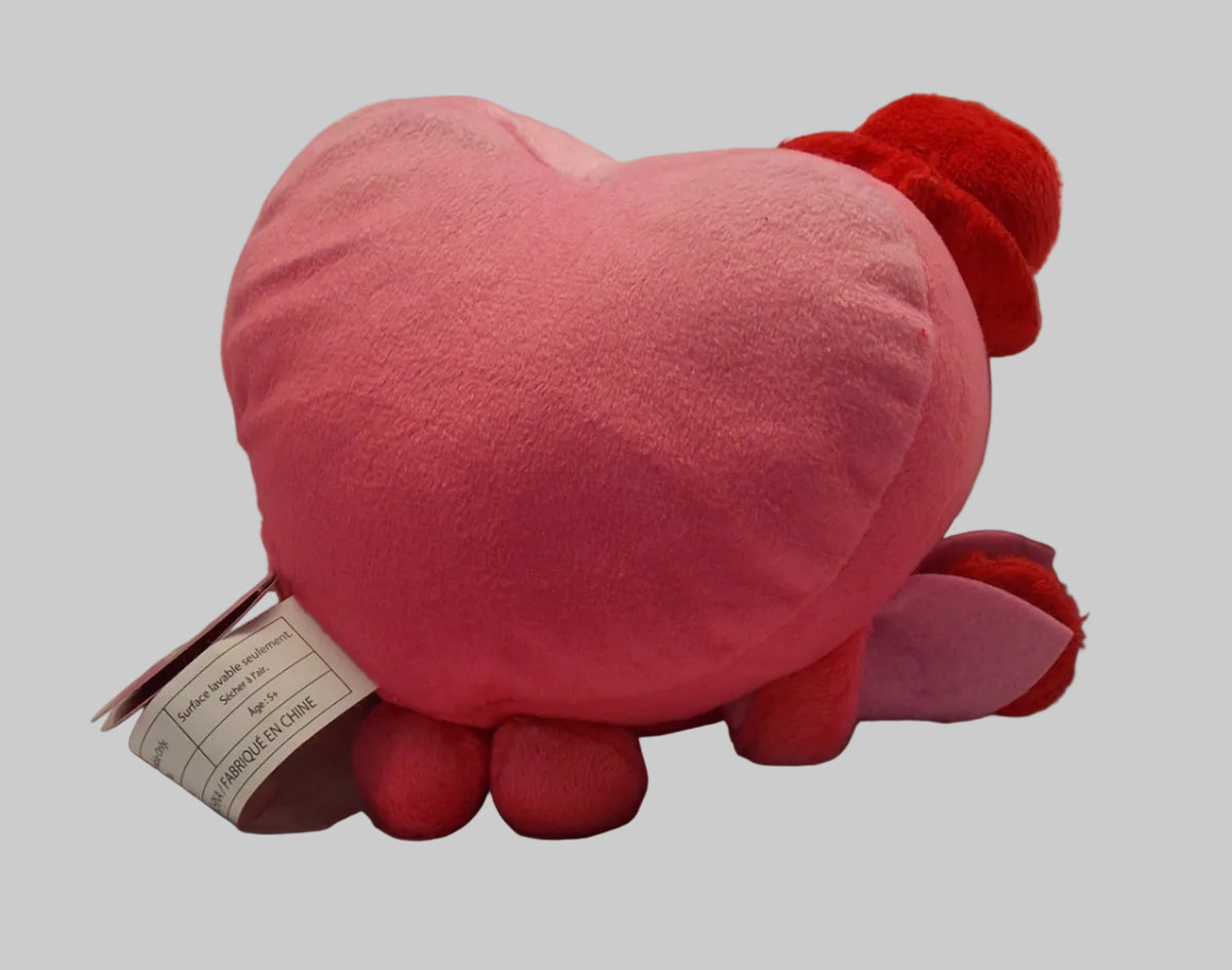 A pink heart-shaped plush pillow with red accents and a white product tag attached, photographed against a wrinkled white fabric background.